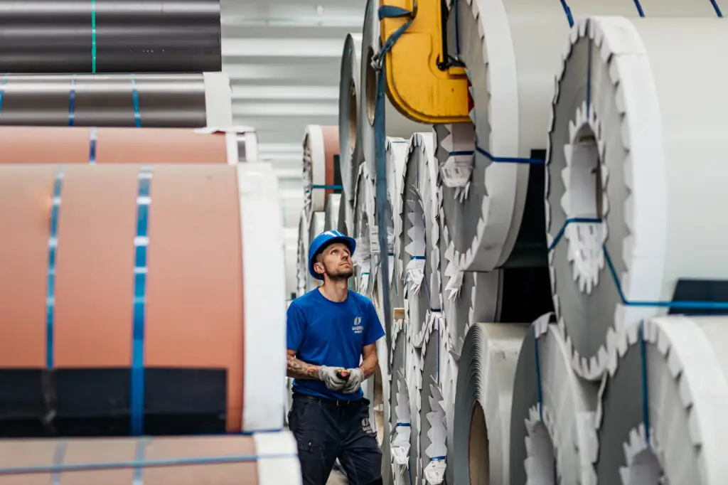 Operaio con casco e guanti osserva bobine metalliche impilate in magazzino industriale, evidenziando lavoro e sicurezza